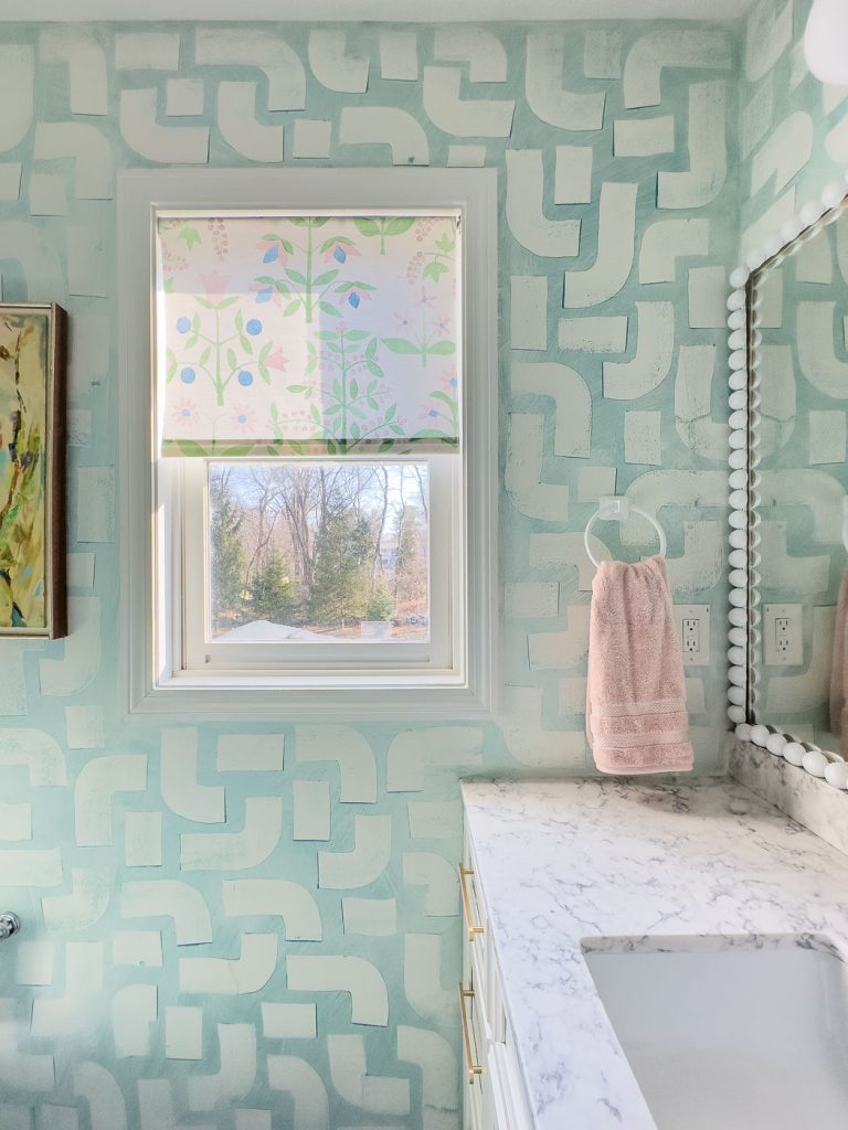 Bathroom with patterned mint-green walls, a window with floral-patterned blinds, and a marble countertop. A pink towel hangs by a circular holder. A large mirror with a white beaded frame reflects soft light.