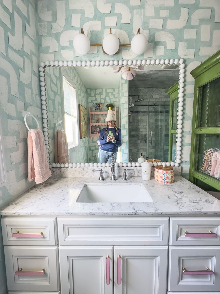 A person takes a mirror selfie in a stylish bathroom with patterned turquoise wallpaper. The vanity has a white marble countertop, pink towel, and decorative items. A large mirror and round light fixtures are above the sink.