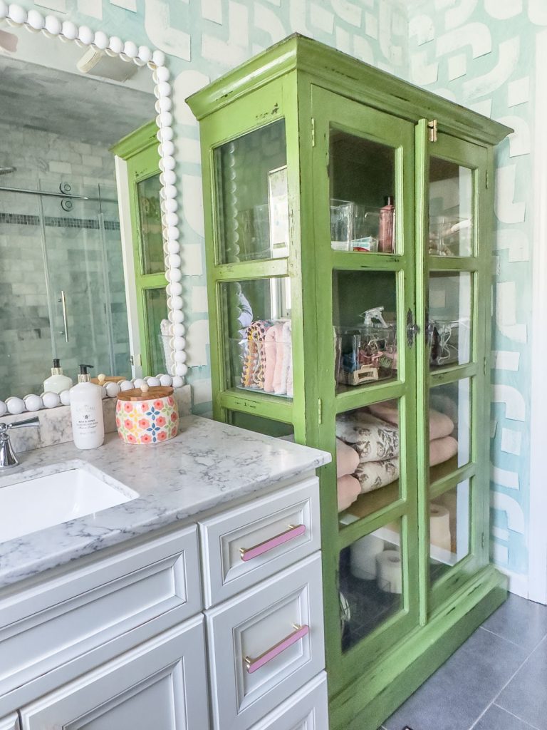 A bathroom features a marble countertop with toiletries and a decorative jar. A green display cabinet with glass doors holds folded towels and various items. The wall has a geometric pattern, and a mirror with a beaded frame is beside the sink.