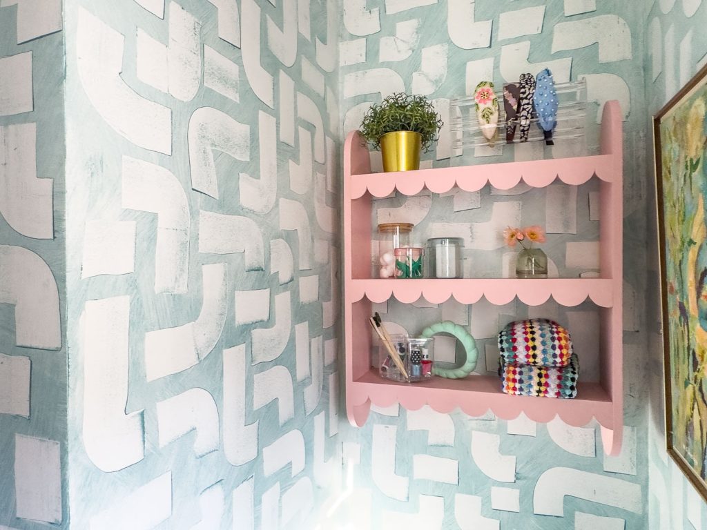 A small corner with a pink wall shelf holding potted greenery, decorative items, and a rolled-up crocheted blanket. The walls are painted with a geometric pattern in white and light blue. An abstract painting is partially visible to the right.