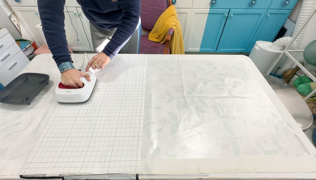 Person using a Cricut heat press on a grid-patterned surface, with a yellow chair and a turquoise cabinet in the background. A roll of transparent film with faint designs is partially visible on the table.