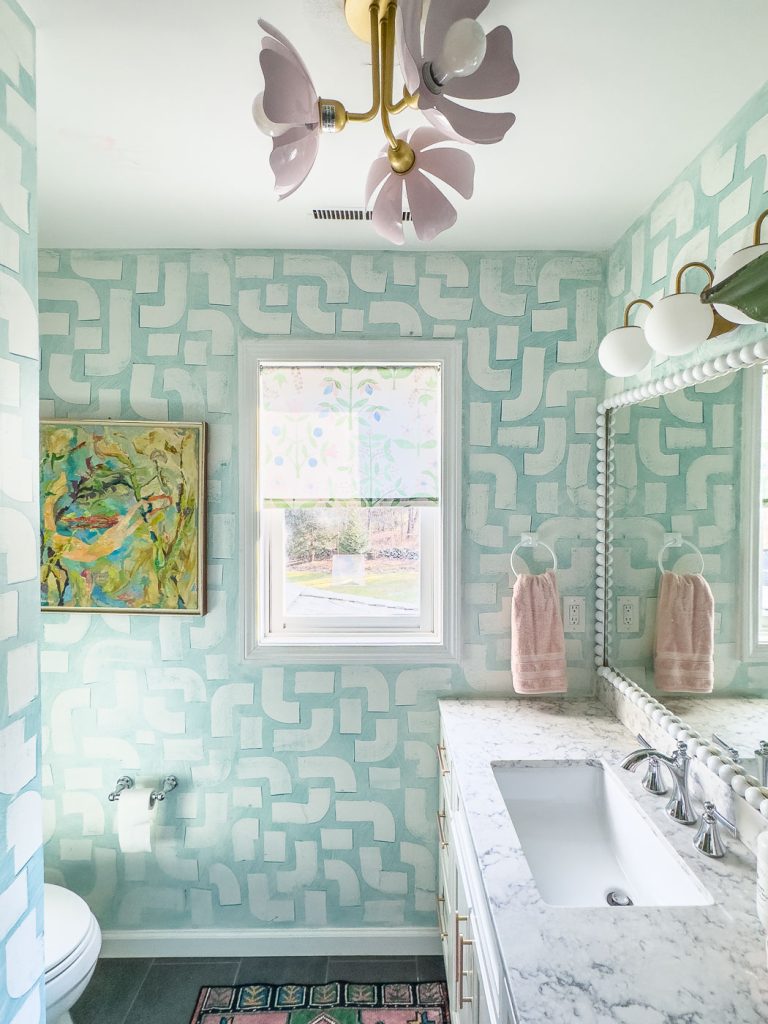 Bathroom with light blue and white patterned walls, white marble countertop, and silver fixtures. A window with floral shades overlooks the room. A colorful abstract painting hangs on the wall. Pink towels and a floral ceiling light fixture add charm.