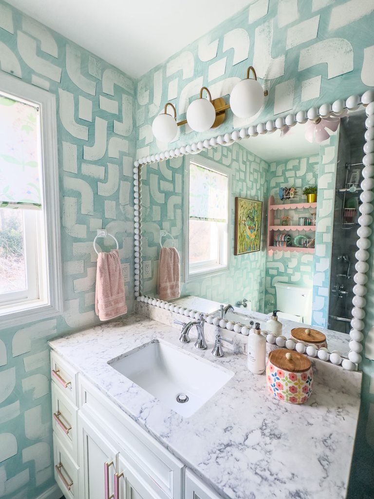 A stylish bathroom with a marble countertop, a large square mirror framed with beads, and modern light fixtures above. The walls have a blue and white geometric pattern. A pink towel hangs on the left, and colorful decor accents the space.