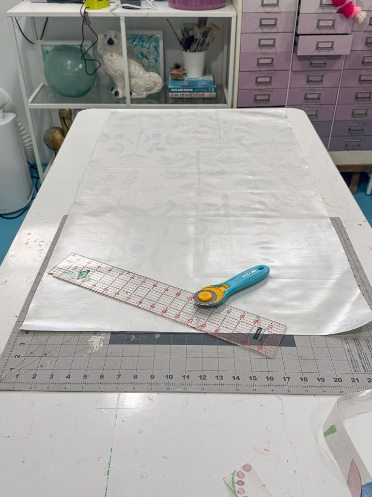 A crafting workspace with a cutting mat on the table, a transparent ruler, and a rotary cutter. Shelves with books and decorative items are in the background, and a pink drawer unit is visible. Various tools are scattered around.