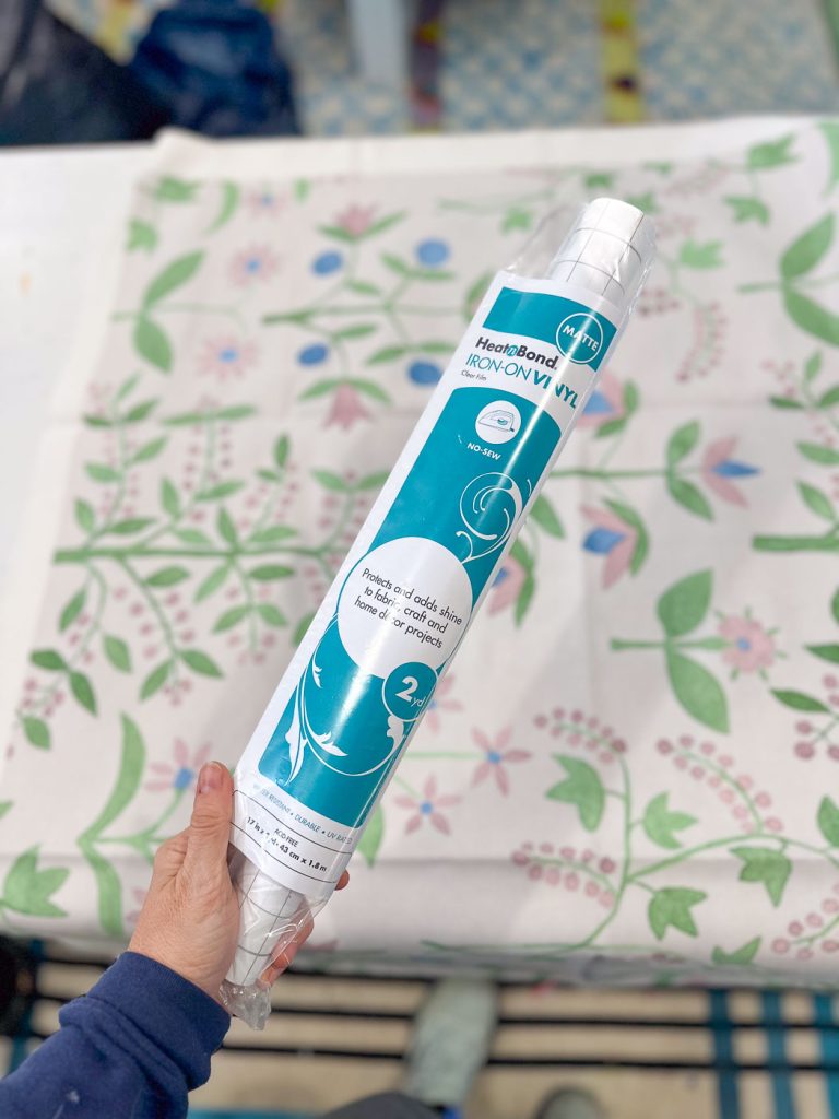 A person holds a roll of HeatnBond Iron-On Vinyl in their hand. The roll is blue and white, featuring product details and instructions. In the background, a partially visible floral-patterned fabric is placed on a table.