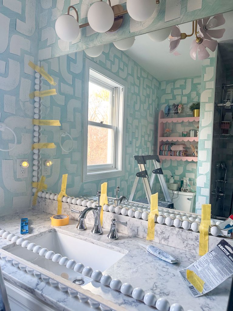 A bathroom with a marble countertop features a large mirror surrounded by yellow tape and decorations. The wall has a blue geometric pattern. A step ladder is visible near shelves displaying various items. Soft overhead lighting illuminates the space.