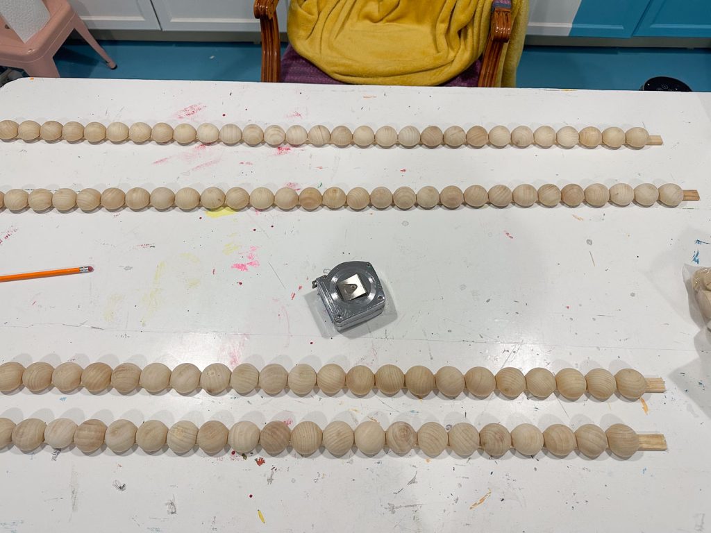 Wooden beads are lined up on dowels on a white table, which has paint splatters. A pencil and a silver pencil sharpener are also on the table. A yellow blanket is draped over a chair in the background.