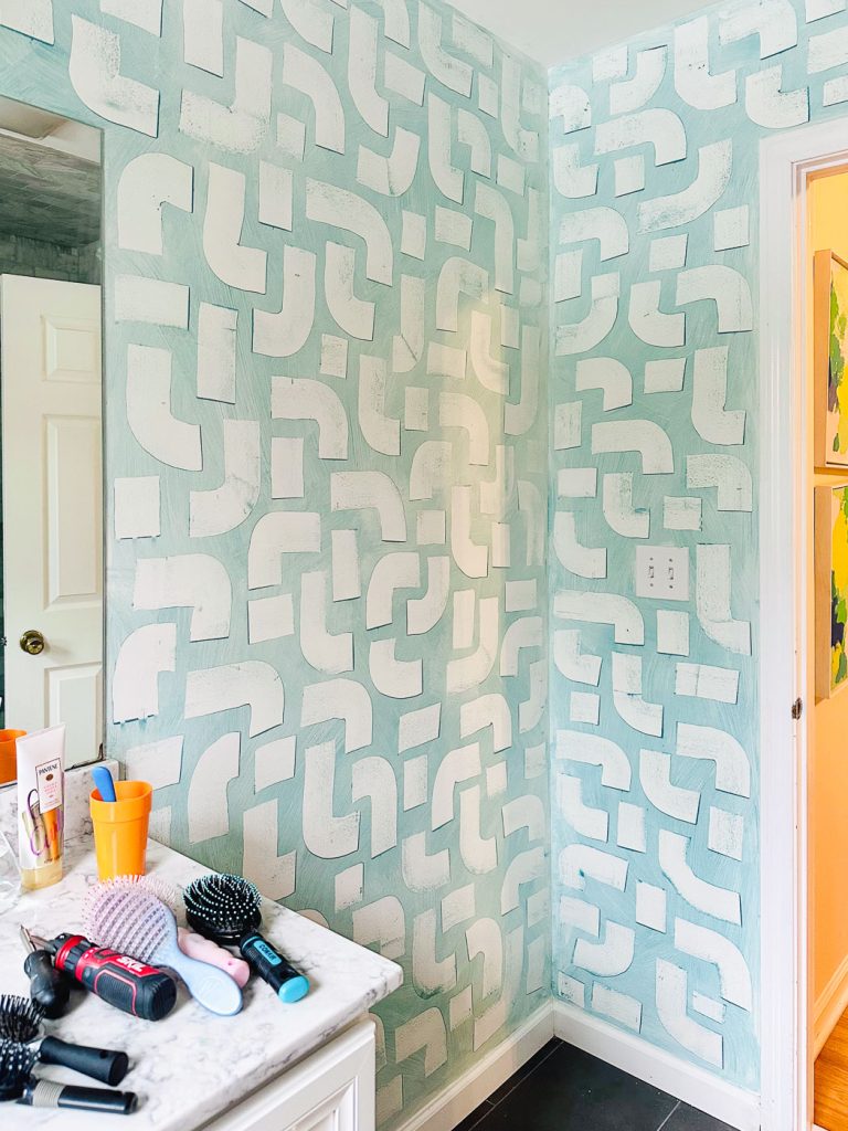 Bathroom with a geometric patterned wall in mint green and white. A countertop holds various hair styling tools and products. A partially open door reveals another room in the background.