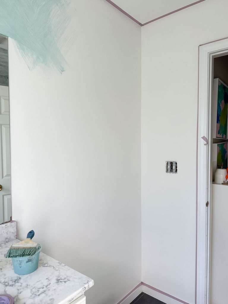 A partially painted room features white walls and trim with light purple painter’s tape. There's a patch of blue-green paint on one wall. A countertop holds painting supplies, including a can of paint and a brush.