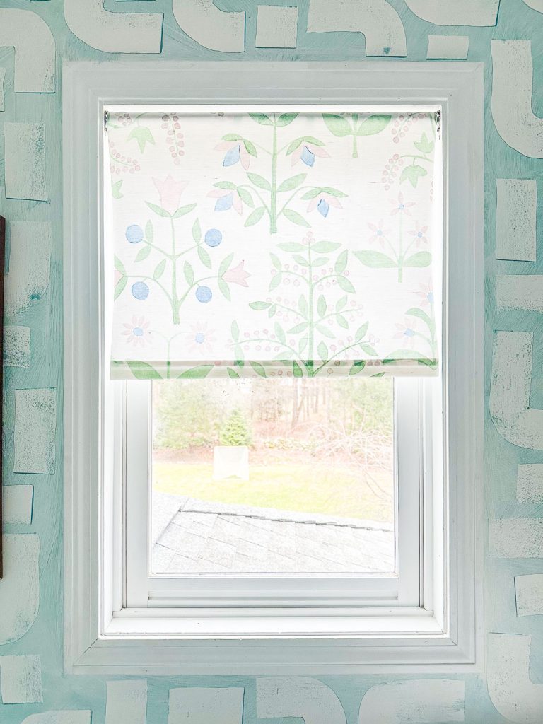 A window with a floral-patterned roller shade featuring light blue and pink flowers is partially rolled down. The wall around the window has a geometric design in shades of light teal and white. Outside, a grass yard is visible.