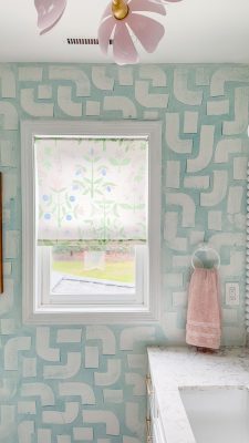 A bathroom with a window showcasing a floral-patterned roller shade complements the light blue geometric walls. A pink towel hangs on a silver ring beside the white countertop and sink, while a pink flower-shaped ceiling light adds charm overhead.
