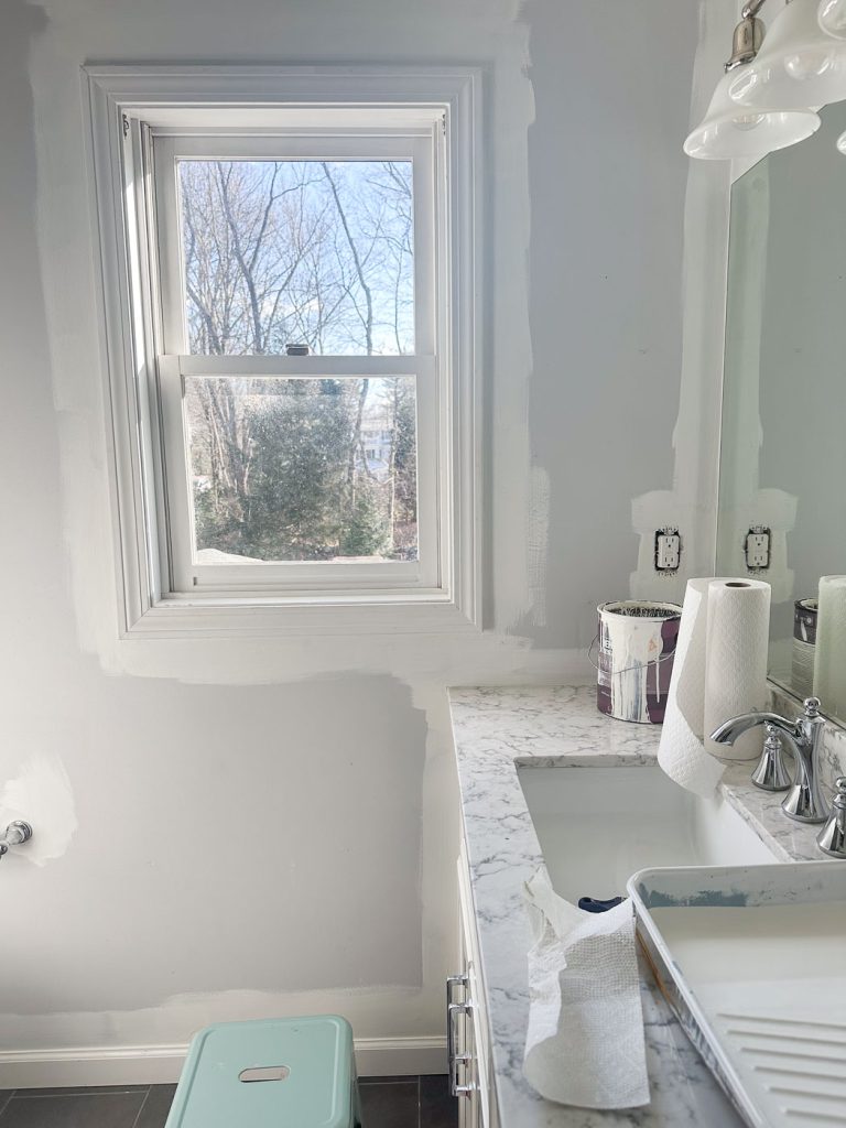 A partially painted bathroom with a white wall around a window. A sink with a marble countertop holds painting supplies, including a roller tray, paper towels, and a paint can. A small stool is on the gray tiled floor. Trees are visible through the window.