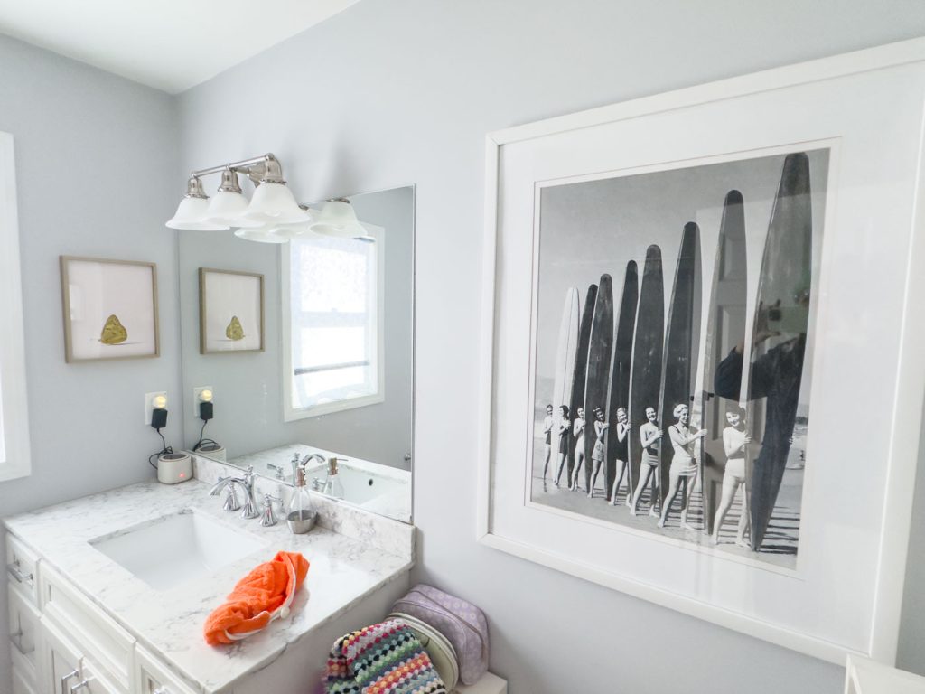 A bathroom with a white vanity and marble countertop. An orange towel is near the sink. There is a framed black-and-white photo of people with surfboards on the wall. Two small, framed images hang above the toilet with a multicolored towel.