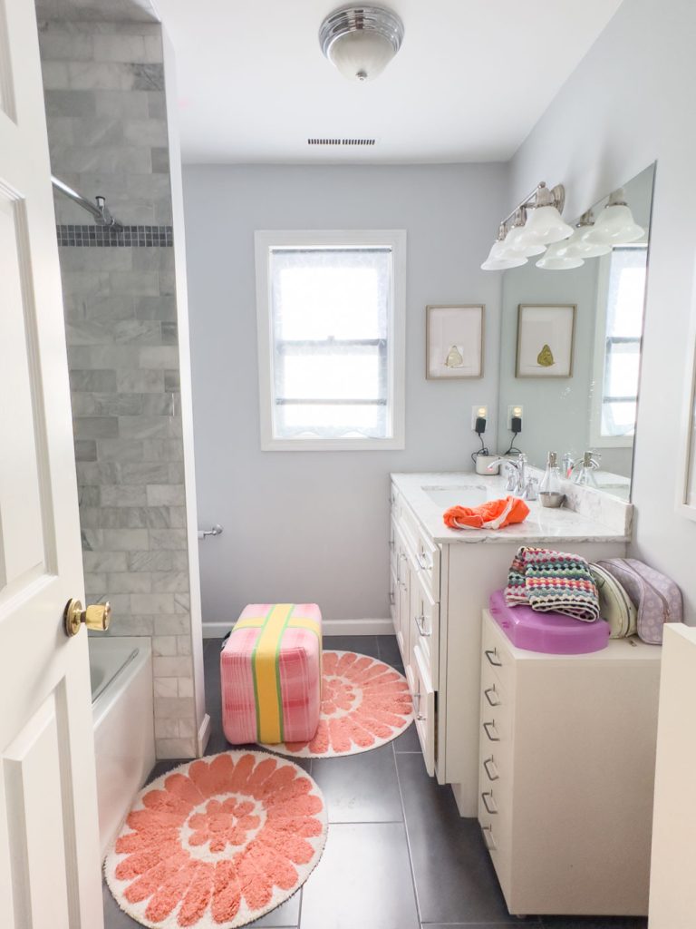 A bright bathroom with a bathtub on the left and a vanity on the right. The countertop holds toiletries and a pink towel. A colorful ottoman and round orange-pink rug are on the floor. Two small framed pictures hang above the toilet.