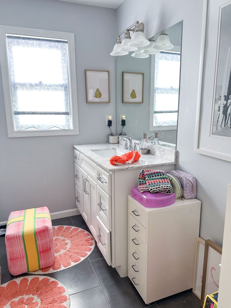 A bathroom with a white vanity, a large mirror, and decorative art on the walls. The counter holds a red hairdryer, a colorful knitted item, and cosmetic bags. A pink and green pouf sits beside a coral and white floral rug on the tile floor.