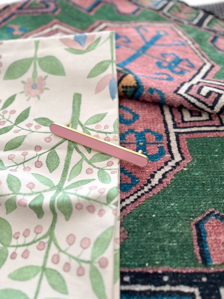 A pink and gold hairpin rests on floral-patterned fabric next to a multicolored geometric carpet. The soft and intricate designs feature green foliage and pink flowers, contrasting with the vivid green, pink, and blue carpet patterns.