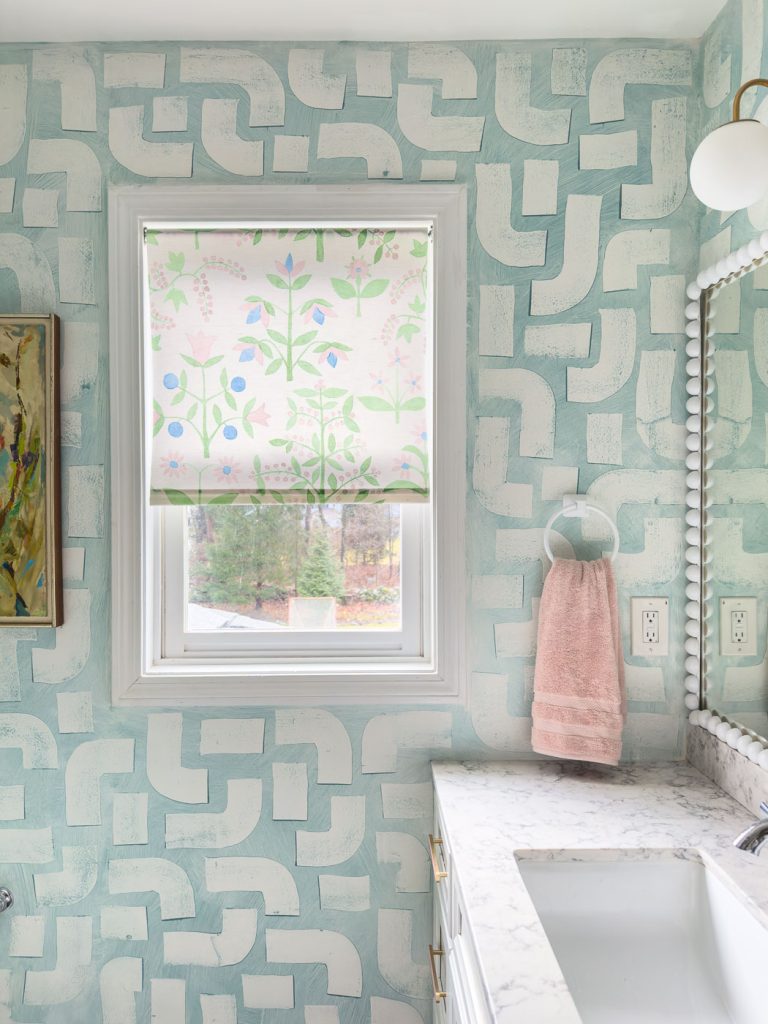 The bathroom showcases blue and white geometric patterned wallpaper, complemented by a floral roller shade that adds a stylish touch. A pink towel hangs nearby, while the marble-patterned countertop and round light fixture enhance the decor above a decorative mirror.