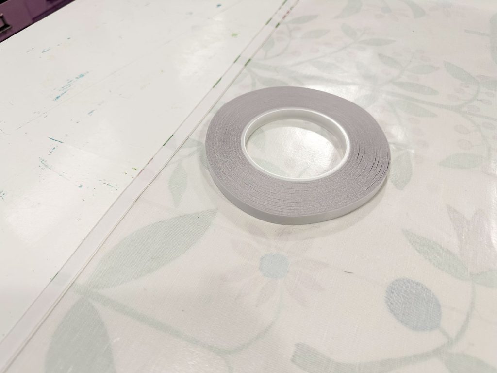 A roll of thin white tape on a light-colored floral-patterned tablecloth. The background includes part of a white desk and some indistinct colored objects.