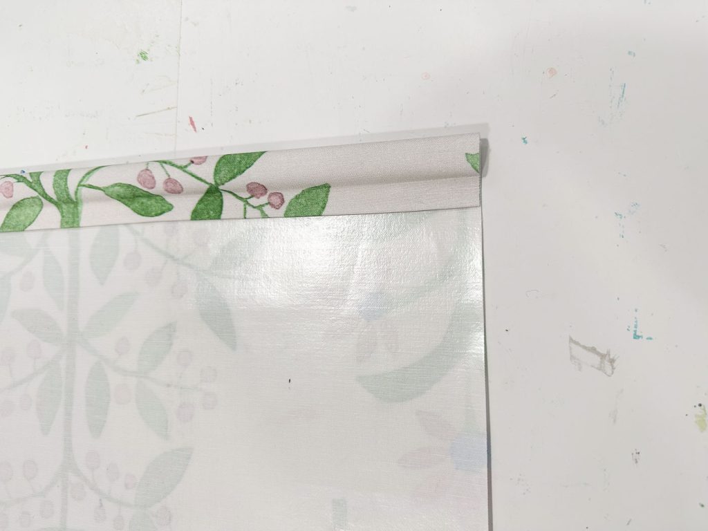 A close-up of a printed floral wallpaper edge on a white surface, featuring green leaves and pink berries. The wallpaper is rolled slightly, revealing the pattern and texture. Marks and smudges are visible on the white background.