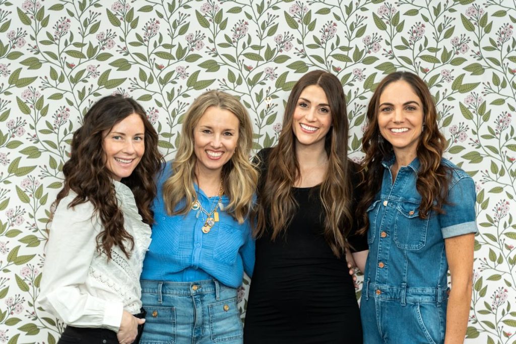 Just 4 Moms Four women are standing in front of a floral wallpaper. They are smiling and dressed in casual attire, including a white blouse, blue top with denim pants, black dress, and denim dress.