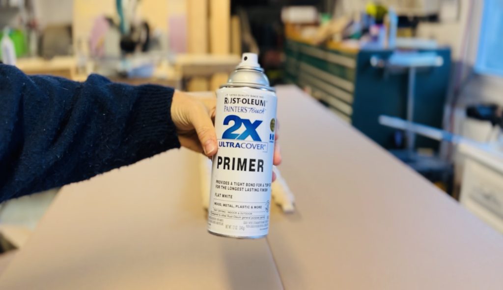 A hand holds a can of Rust-Oleum Painter's Touch 2X Ultra Cover Primer in a workshop. The background includes blurred tools and a workbench, suggesting a DIY or painting project setting.