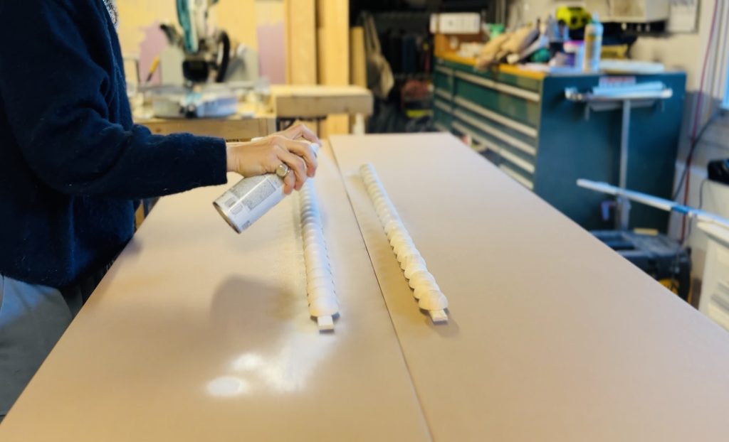 A person is spray-painting two long objects placed on a table in a workshop. The background shows various tools and equipment. The scene suggests a woodworking or crafting project in progress.