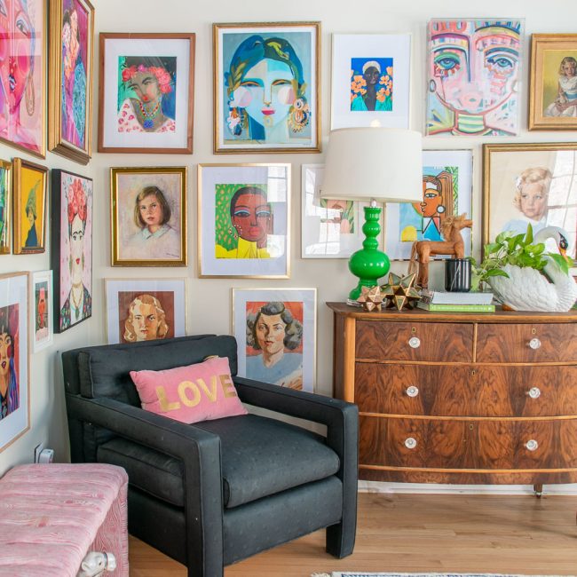 A cozy living room with a black armchair and a cushioned stool. The walls are adorned with vibrant portraits in various styles and frames. A wooden dresser holds a green lamp and decorative pieces, and there's a fluffy stool nearby.