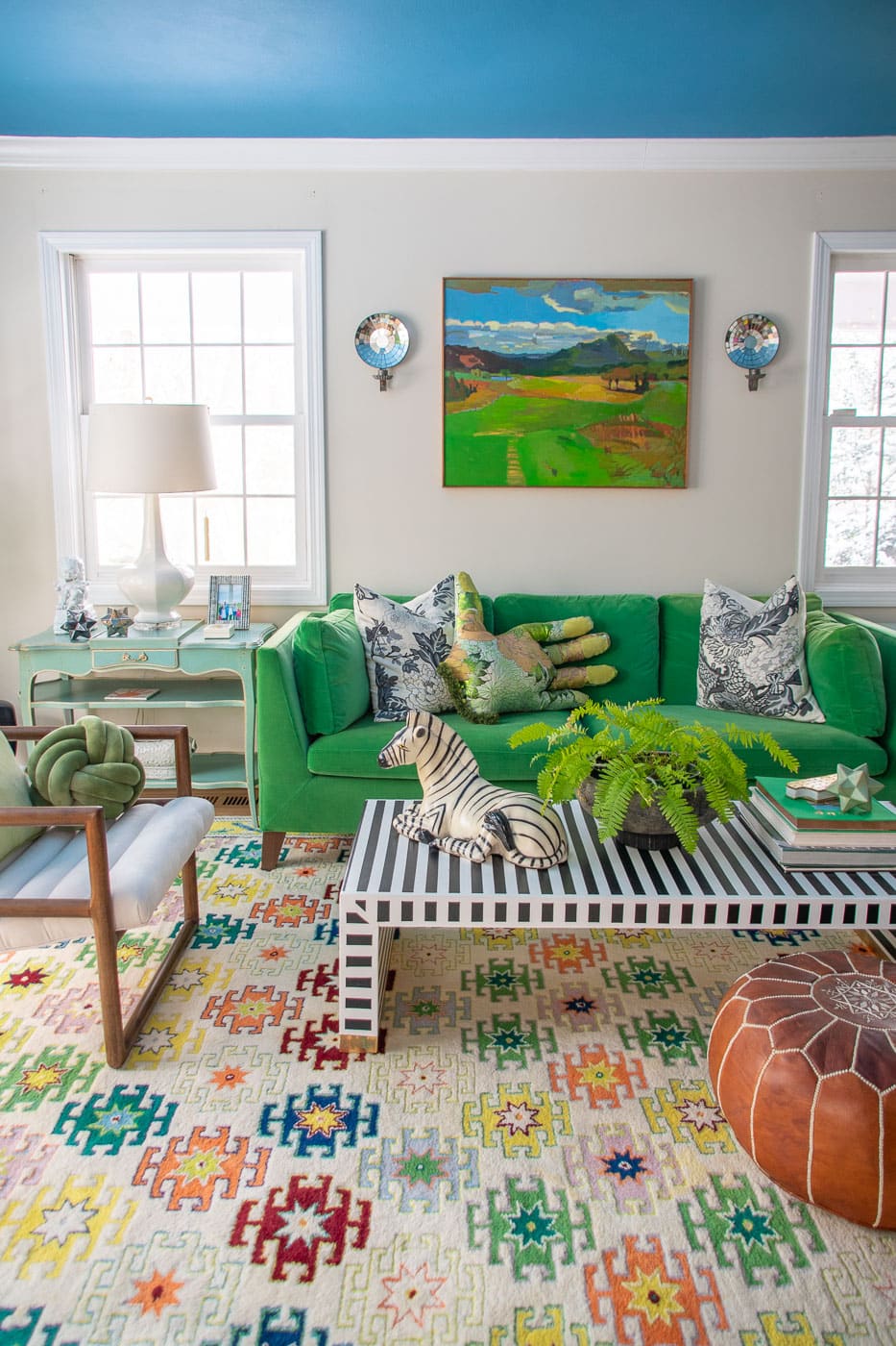 A vibrant living room with a green sofa adorned with patterned pillows, a striped black and white coffee table holding decor items, a colorful patterned rug, artwork on the wall, and a large window letting in natural light.