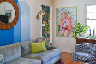 Living room with a blue and gray sofa, green and patterned cushions, large colorful painting on the wall, round wooden mirror, and a plant on a wooden side table. The walls feature blue accents and a decorative lamp.