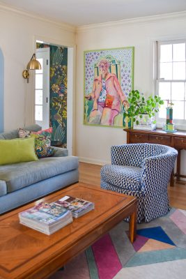 Colorful living room with a blue couch, patterned blue and white armchair, wooden coffee table with magazines, abstract rug, and vibrant artwork on the wall. A gold wall lamp and potted plant add decorative touches. Natural light fills the room.