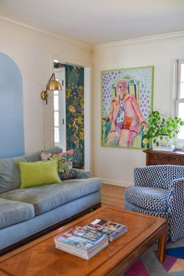 A living room with a light blue sofa, a patterned blue and white chair, and a wooden coffee table with magazines. A large colorful portrait painting is on the wall. There's a gold wall lamp and a green plant on a wooden sideboard.