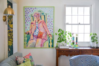 Living room with a colorful portrait of a person seated in a chair, wearing vibrant clothing and holding a snack. The room features a green plant on a wooden table, patterned cushions on a sofa, and a large window letting in natural light.