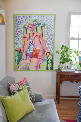 A colorful portrait painting of a person with exaggerated features sits on a wall above a couch. The room also has a small table with a potted plant and a window letting in natural light. Various colorful cushions are on the couch.