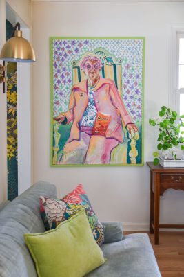 A colorful painting of a person wearing a pink suit and seated on a green chair hangs on the wall. They hold a bag of Cheetos. The room has a gray couch with vibrant pillows and a wooden side table with a potted plant.