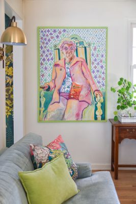 Colorful interior with a large vibrant painting on the wall depicting a person in pink attire holding a book. A light blue couch with multicolored pillows is beneath, and a small wooden table with a plant is nearby.