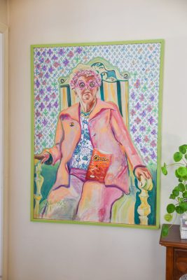 A colorful painting of a person wearing a pink coat and round glasses, seated on a striped chair. The person holds a bag of Cheetos, with vibrant wallpaper in the background and a plant visible in the foreground.