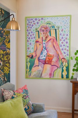 A colorful painting of a person in a pink outfit sitting on an ornate chair, holding a Cheez-Its box against a patterned wall. The room features a floral-patterned chair, a gold lamp, and green leafy plants decorating the space.