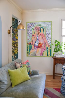 A cozy living room with a light blue sofa, colorful pillows, and a vibrant painting of a person seated in a chair. A green plant sits on a wooden table by a window. A modern gold floor lamp arches over the sofa.