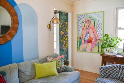 A cozy living room with a blue and white sofa, colorful throw pillows, and a gold floor lamp. A large, vibrant painting hangs on the wall next to a window and potted plant. The room has a mix of blue tones and a wooden side table.