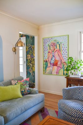A cozy living room with a light blue sofa, green and patterned cushions, and a blue patterned chair. A colorful painting of a person on a deck chair is on the wall. A wooden table with a leafy plant sits beside the chair. Bright wallpaper peeks from a doorway.