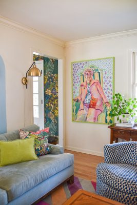 A cozy living room with a blue couch, green and multicolored pillows, floral and geometric-patterned decor, and a large colorful painting of a person on the wall. A leafy plant sits on a wooden cabinet beside a patterned chair.