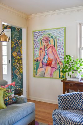 A vibrant room features a large, colorful painting of a person seated, with an abstract and playful style. The walls are adorned with floral and bird-patterned wallpaper. A leafy plant sits on a wooden side table near a patterned armchair and couch.