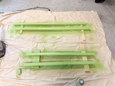 Two wooden frames lie on a drop cloth, spray-painted in light green, with paint cans nearby. The floor is speckled, suggesting a workshop or garage setting.