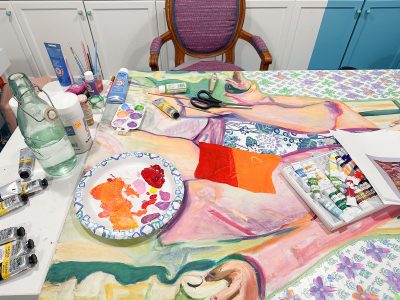 A table cluttered with art supplies, including paint tubes, brushes, scissors, and a paper plate with paint. A colorful painting in progress covers most of the tabletop. A pink-cushioned chair is positioned behind the table.