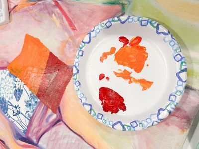 A paper plate with blobs of red and orange paint sits on a painted surface. To the left, there's a piece of red and orange painted paper. The background includes soft pastel-colored shapes.