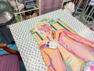 A colorful painting of a person in an orange coat and pink glasses lies on a patterned table. A small paint palette rests on the artwork. Surrounding items include a pink cabinet and various art supplies in a cluttered studio setting.