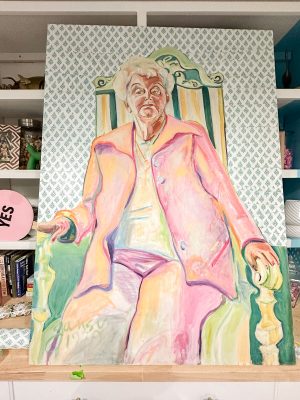 A colorful painting of an elderly person sitting in a green-trimmed chair. They’re wearing a pink coat over a white outfit. The background features a patterned wall, and the painting is placed against a shelf with various items.