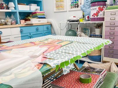 A vibrant craft room with a desk covered in a partially completed jigsaw puzzle. Bright blue cabinets, stacked books, and a colorful mug create a lively backdrop. Various crafting supplies are organized neatly across the shelves.
