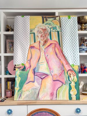 A colorful painting of an older woman seated, wearing a pink suit. The artwork is taped with patterned paper on the sides and is set against a shelf filled with books, jars, and various decorations.