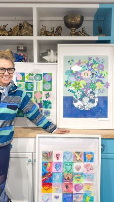 A person in a striped sweater stands beside three colorful framed artworks. The artworks feature geometric shapes, a vase with flowers, and a grid of hearts. Decorative items are displayed on shelves behind them.